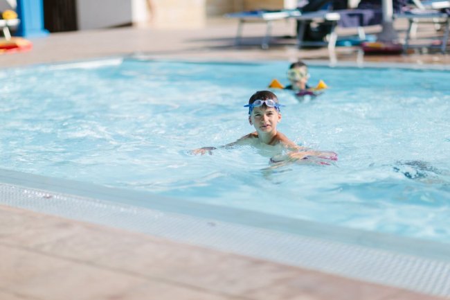 Un bambino in piscina con occhialini e braccioli, altri nuotatori sullo sfondo.