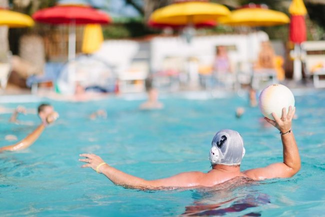 Un uomo in piscina gioca a pallanuoto, sfondo con ombrelloni colorati.