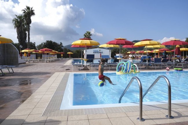 Una piscina con ombrelloni gialli e rossi, una persona siede sul bordo. Palmi sullo sfondo.