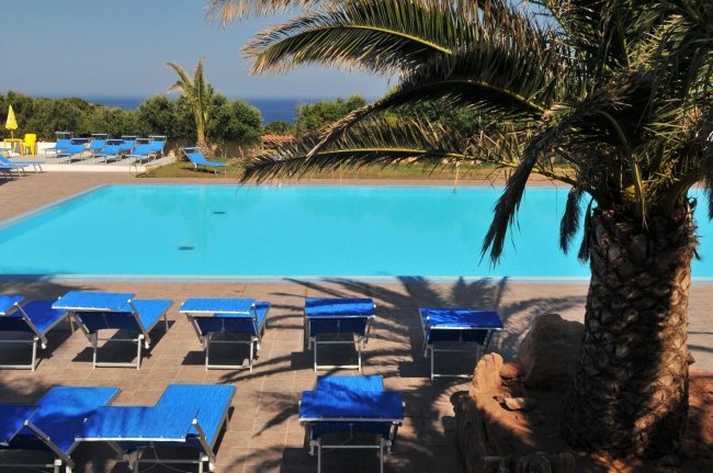 Piscina con lettini blu, circondata da palme, vista mare all'orizzonte.