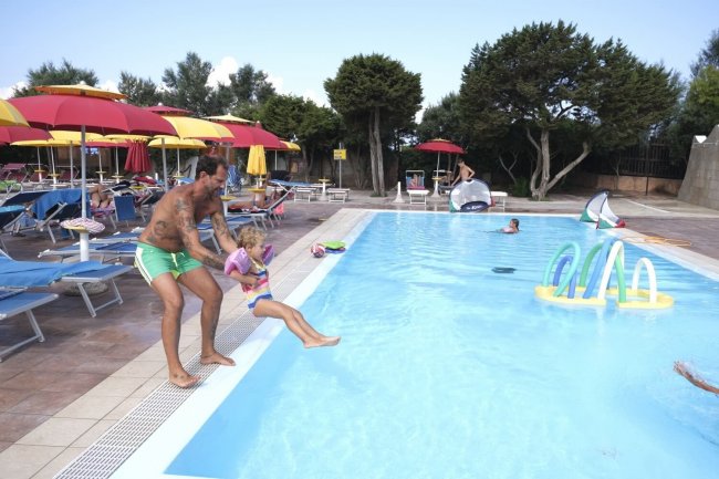 Papà lancia una bambina in piscina; ombrelloni colorati intorno.