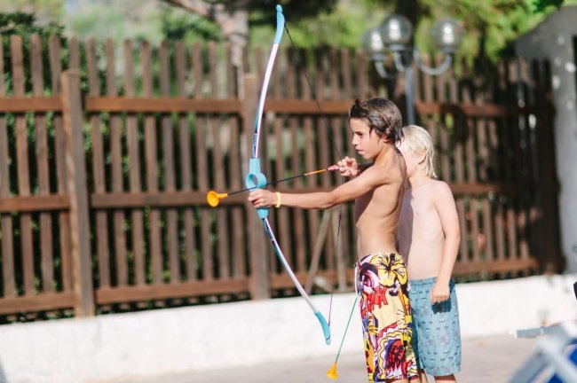 Due bambini giocano con arco e freccia giocattolo, vicino a una staccionata di legno.