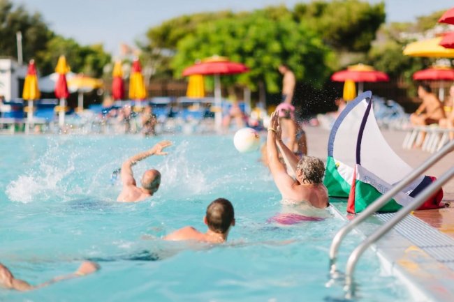 Persone che giocano a pallavolo in piscina con ombrelloni colorati sullo sfondo.