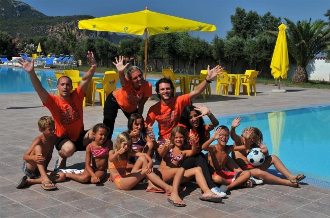Gruppo di bambini e adulti sorridenti vicino a una piscina sotto ombrelloni gialli.
