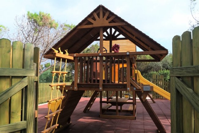 Parco giochi in legno con scivolo giallo e area arrampicata, circondato da recinzione.