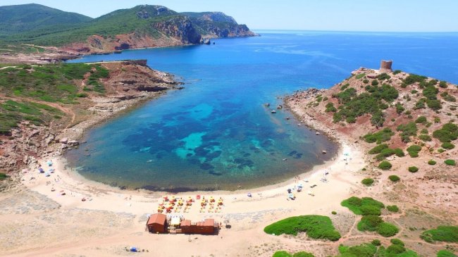 Spiaggia a forma di mezzaluna con mare azzurro e torre su un promontorio.