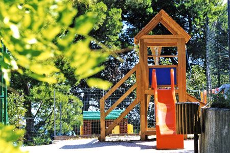 Parco giochi con scivolo arancione e struttura in legno circondato da alberi verdi.