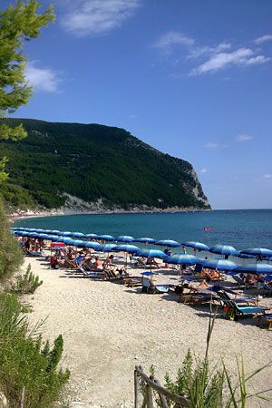 Spiaggia con ombrelloni blu, mare e una montagna verde sullo sfondo.