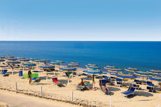 Spiaggia con lettini blu e ombrelloni gialli e blu su sabbia chiara.