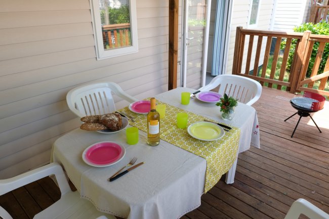 Tavolo di legno apparecchiato con piatti, bicchieri colorati, pane e bottiglia di vino su veranda.