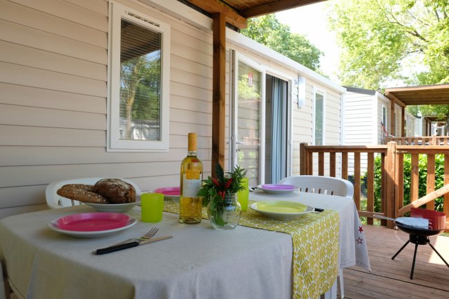 Tavolo all'aperto con stoviglie colorate, pane e vino. Mobile home sullo sfondo.