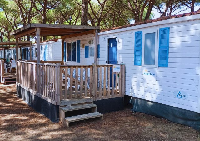 Una casa mobile bianca con persiane blu, veranda in legno e scalini, immersa in un'area alberata.
