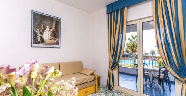 Salotto con divano beige, tende blu e gialle. Finestra aperta su balcone con vista piscina.