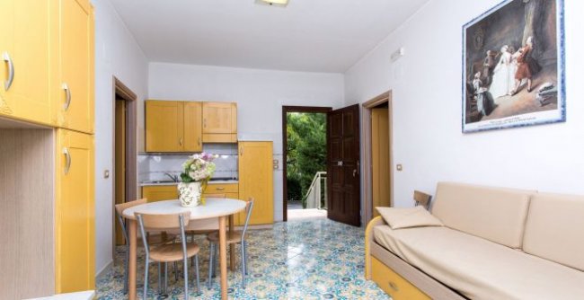 Cucina-soggiorno con mobili gialli, tavolo rotondo, divano beige. Porta aperta su area verde.