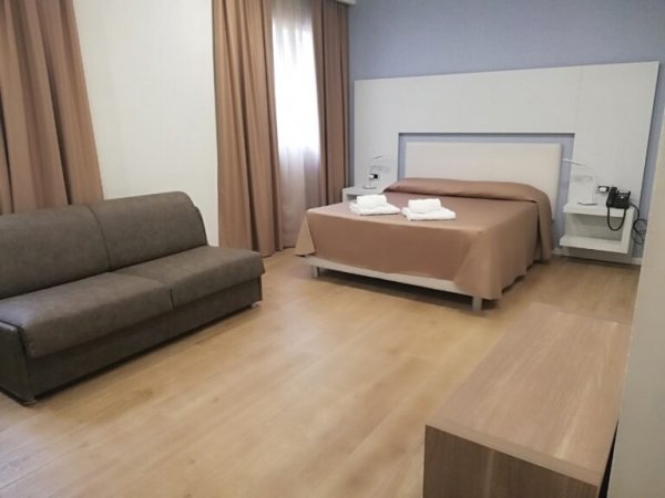 Camera d'albergo con letto matrimoniale, divano grigio e finestra con tende.