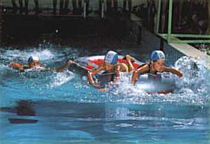 Bambini in gara in piscina con cuffie e palette, schizzano mentre nuotano.