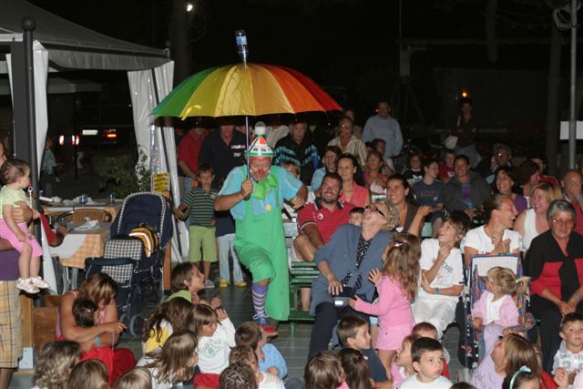 Un clown con un ombrello colorato intrattiene un pubblico di bambini e adulti.