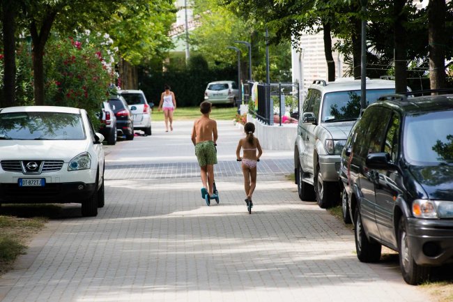 Bambini corrono su un vialetto con auto parcheggiate.