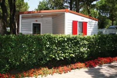 Un bungalow bianco con persiane rosse, circondato da una siepe e bordure di fiori.