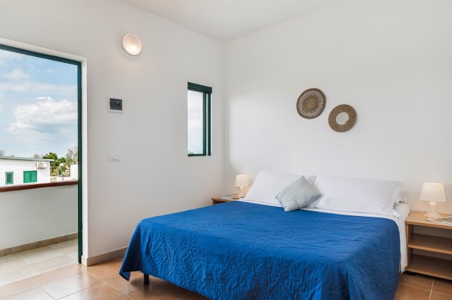 Camera da letto con letto matrimoniale blu, lampade su comodini e porta aperta su balcone.