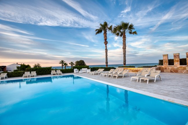 Piscina con lettini e palme, affacciata sul mare al tramonto. Cielo con nubi sfumate.
