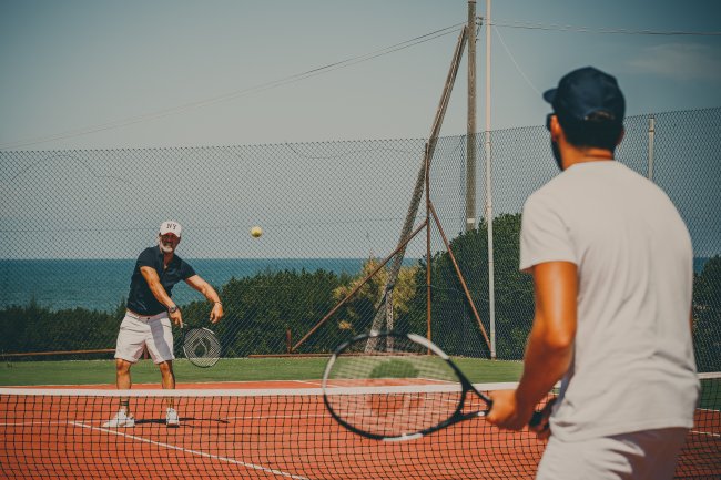 Due persone che giocano a tennis su un campo all'aperto vicino al mare, con una rete di recinzione.