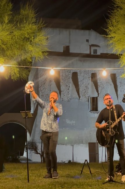 Due musicisti si esibiscono all'aperto con chitarra e tamburello, illuminati da luci sopra di loro.