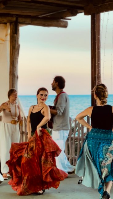 Donne danzano con gonne colorate vicino al mare mentre un uomo suona la chitarra al tramonto.