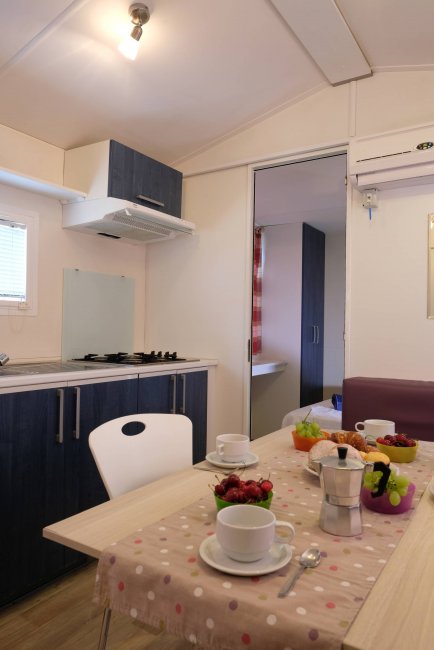 Tavolo da pranzo apparecchiato in cucina con frutta e caffettiera, porta aperta su camera da letto.